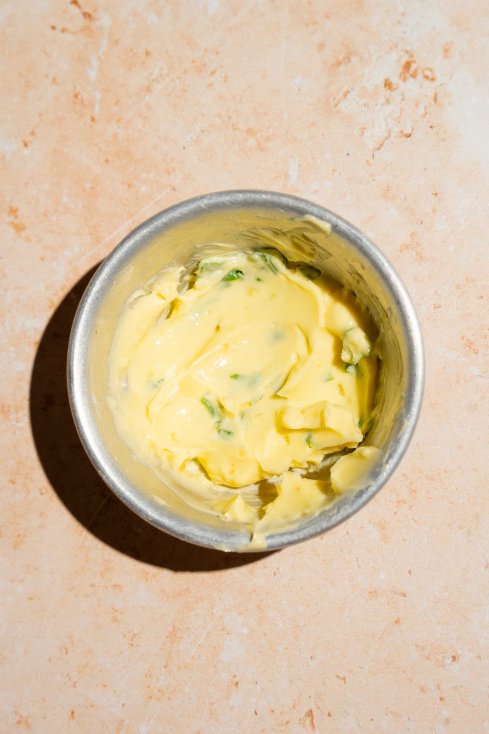 A small bowl of butter mixed iwth fresh herbs like chives and seasonings. The bowl is on a tan counter.