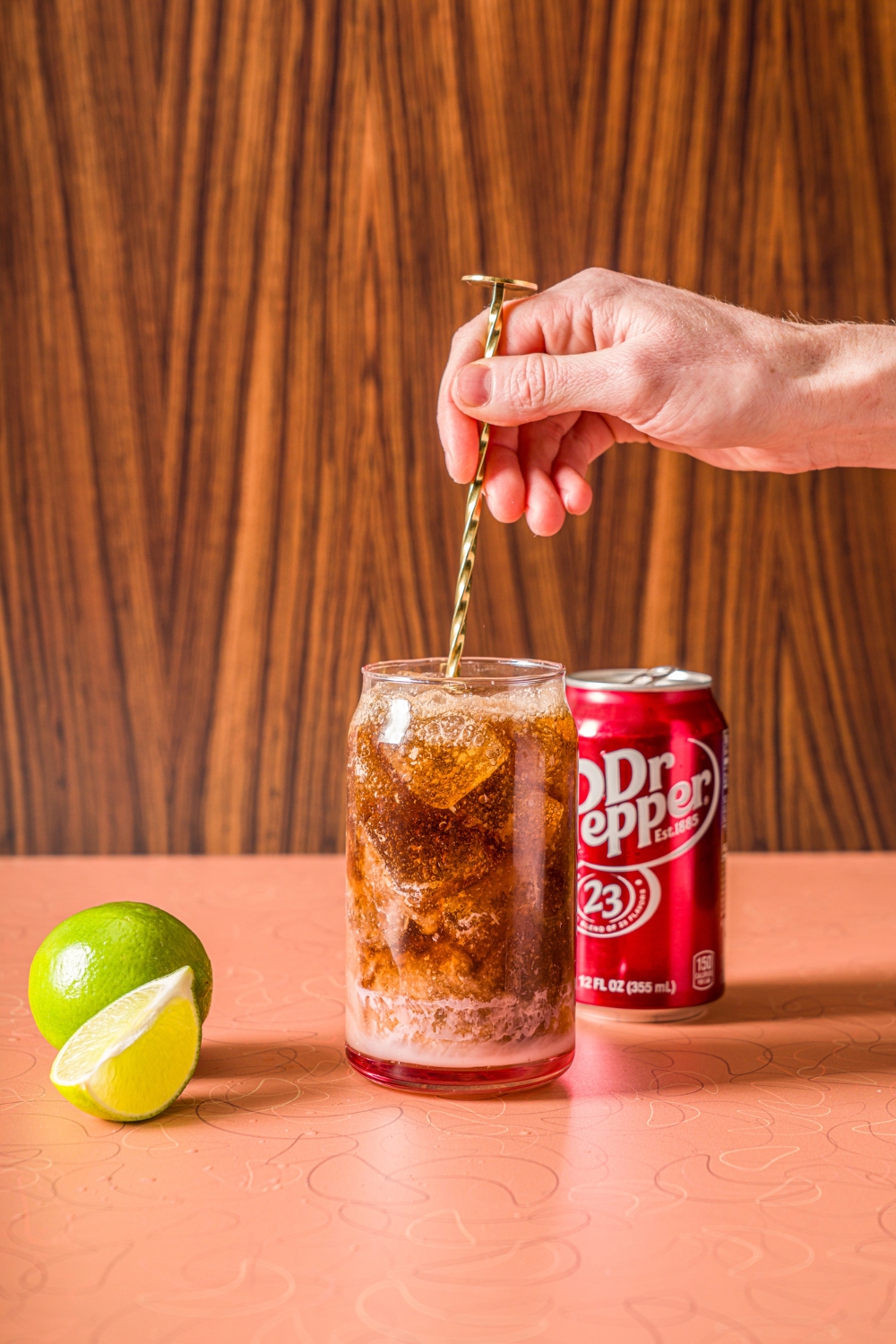 A glass of ice with a hand stirring Dr.Pepper, coconut cream, and vanilla syrup with a stirrer. The glass is on a counter with a can of Dr. Pepper and limes.