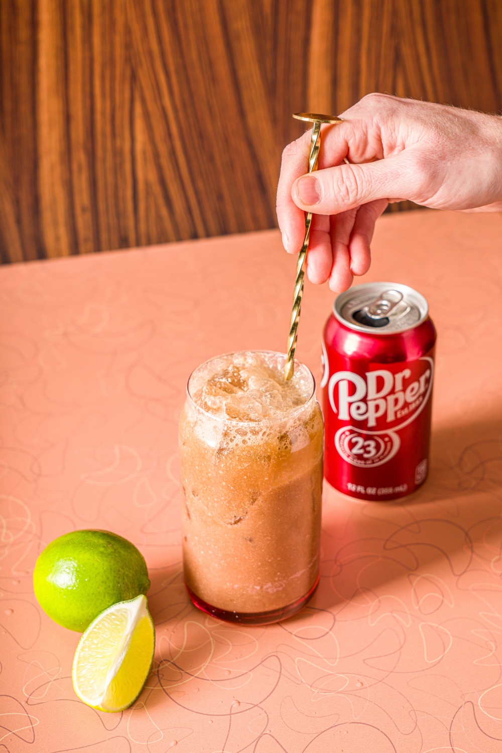 A glass of ice with a hand stirring Dr.Pepper, coconut cream, and vanilla syrup with a stirrer. The glass is on a counter with a can of Dr. Pepper and limes.