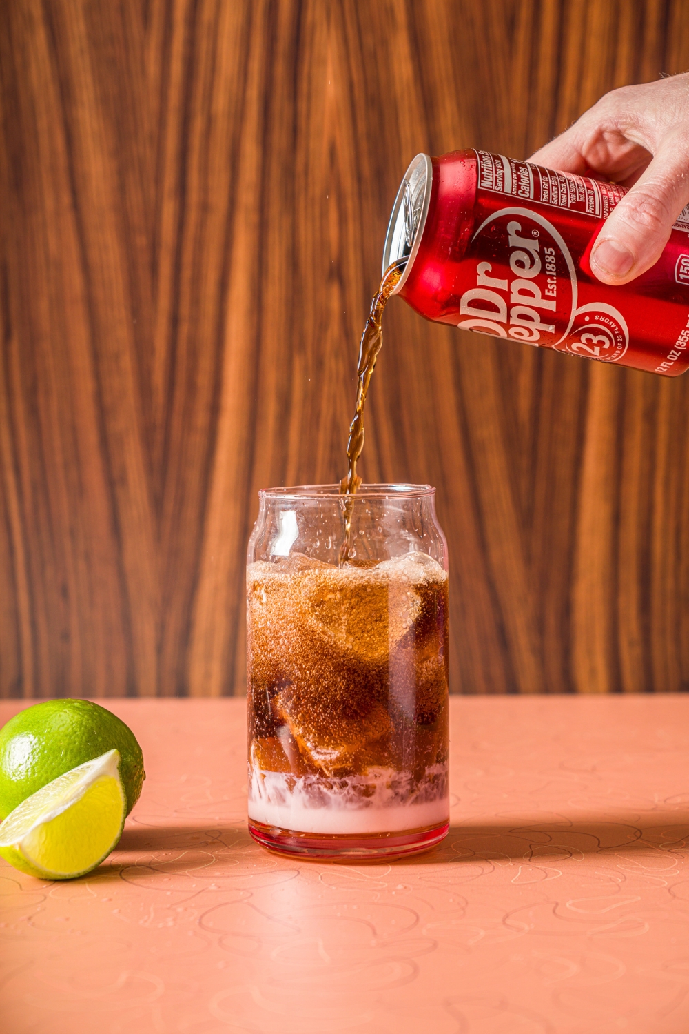 A glass of ice with coconut cream, vanilla syrup, and a can of Dr. Pepper being poured into the glass. The glass is on a counter with limes.