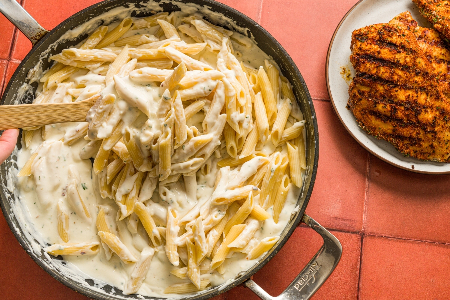 A skillet with penne pasta tossed in alfredo sauce with a wooden spoon. The skillet is on a red counter with a plate of blackened chicken.
