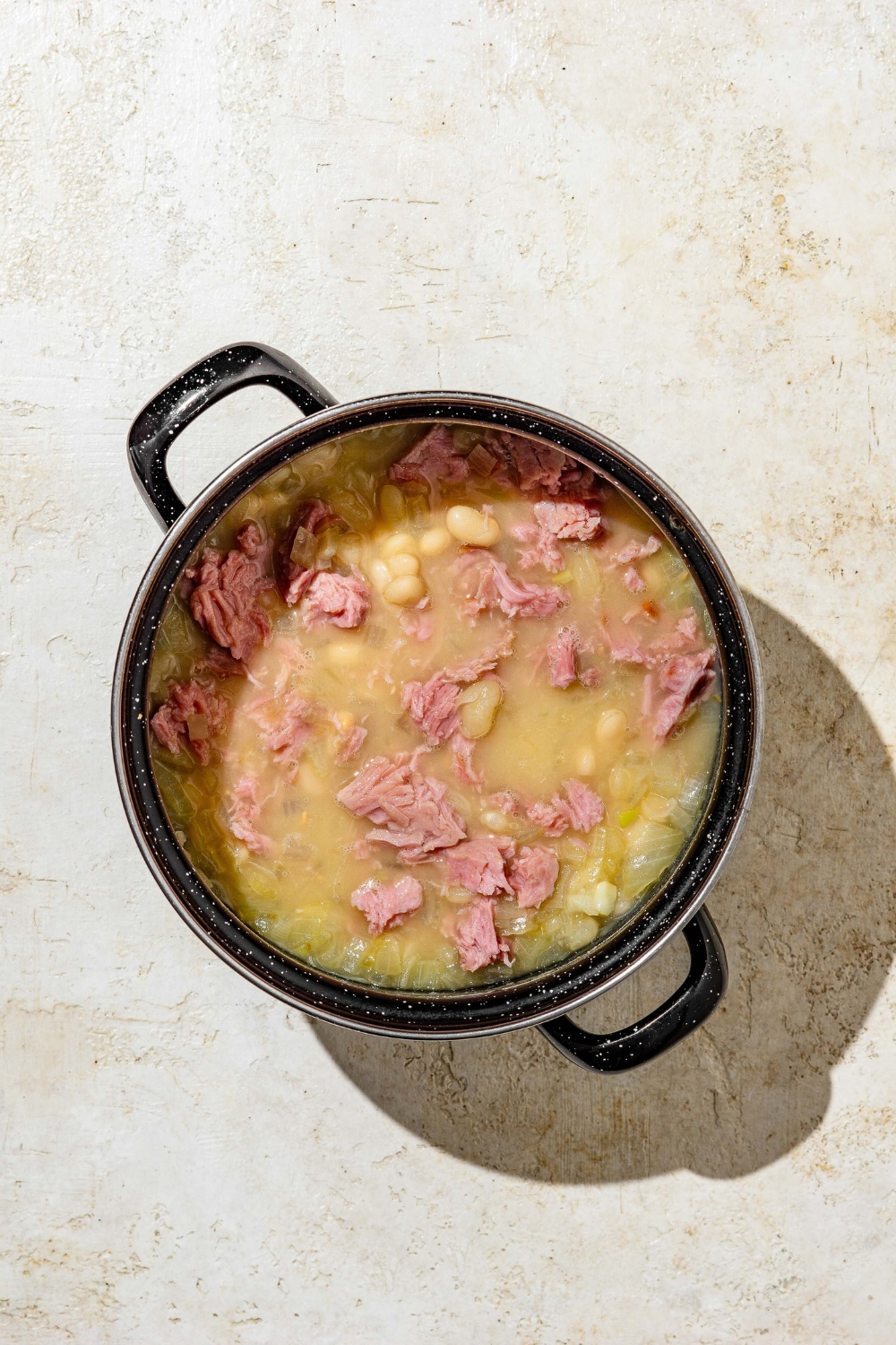 A stock pot with cooked senate bean soup topped with ham. The pot is on a tan counter.