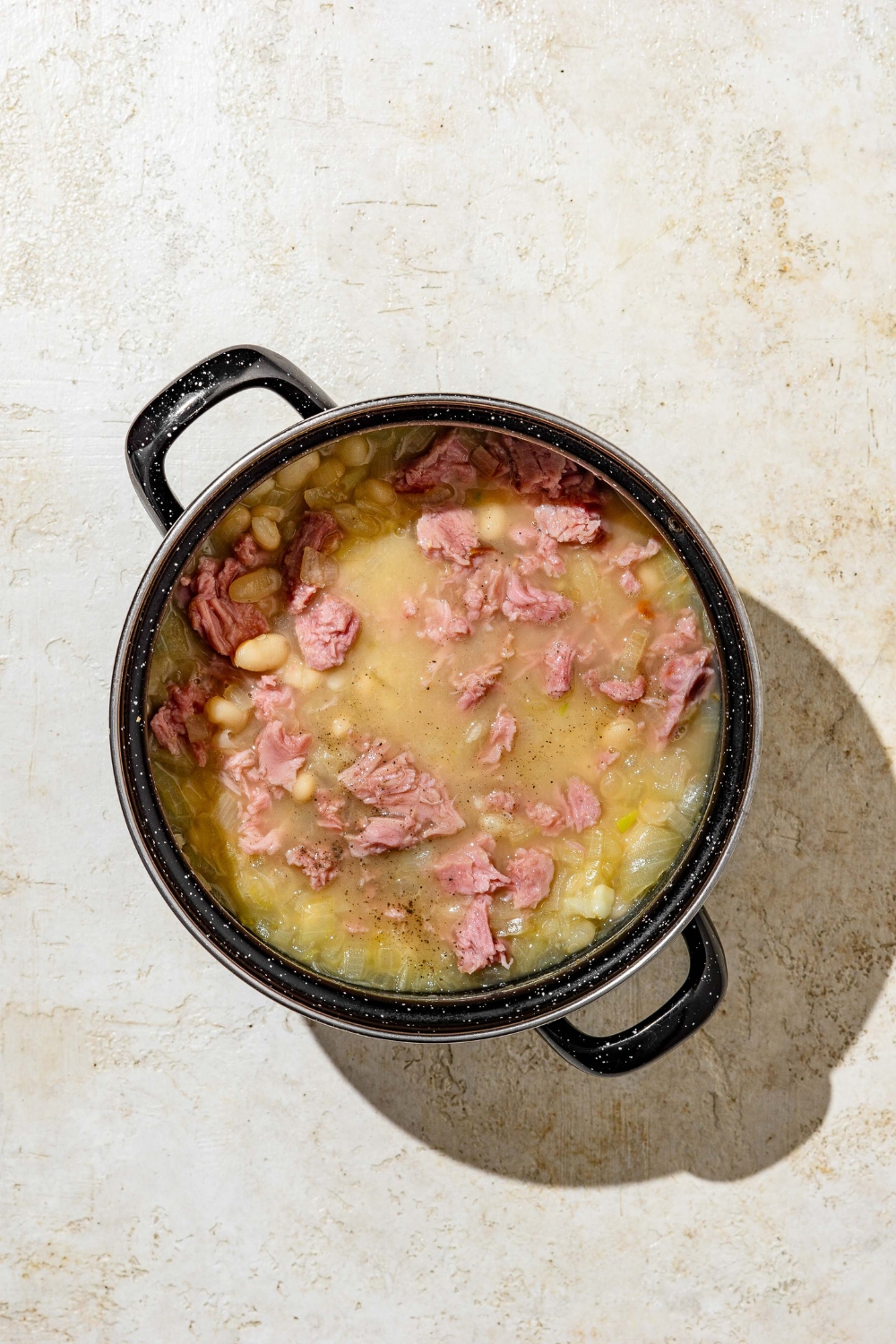 A stock pot with cooked senate bean soup topped with ham. The pot is on a tan counter.