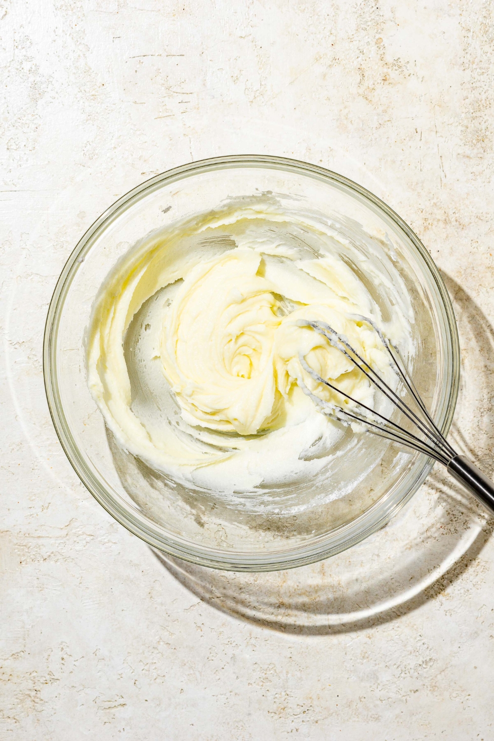 A white bowl with a whisk mixing ingredients to make white Texas sheet cake icing including powdered sugar and butter. The bowl is on a white counter.