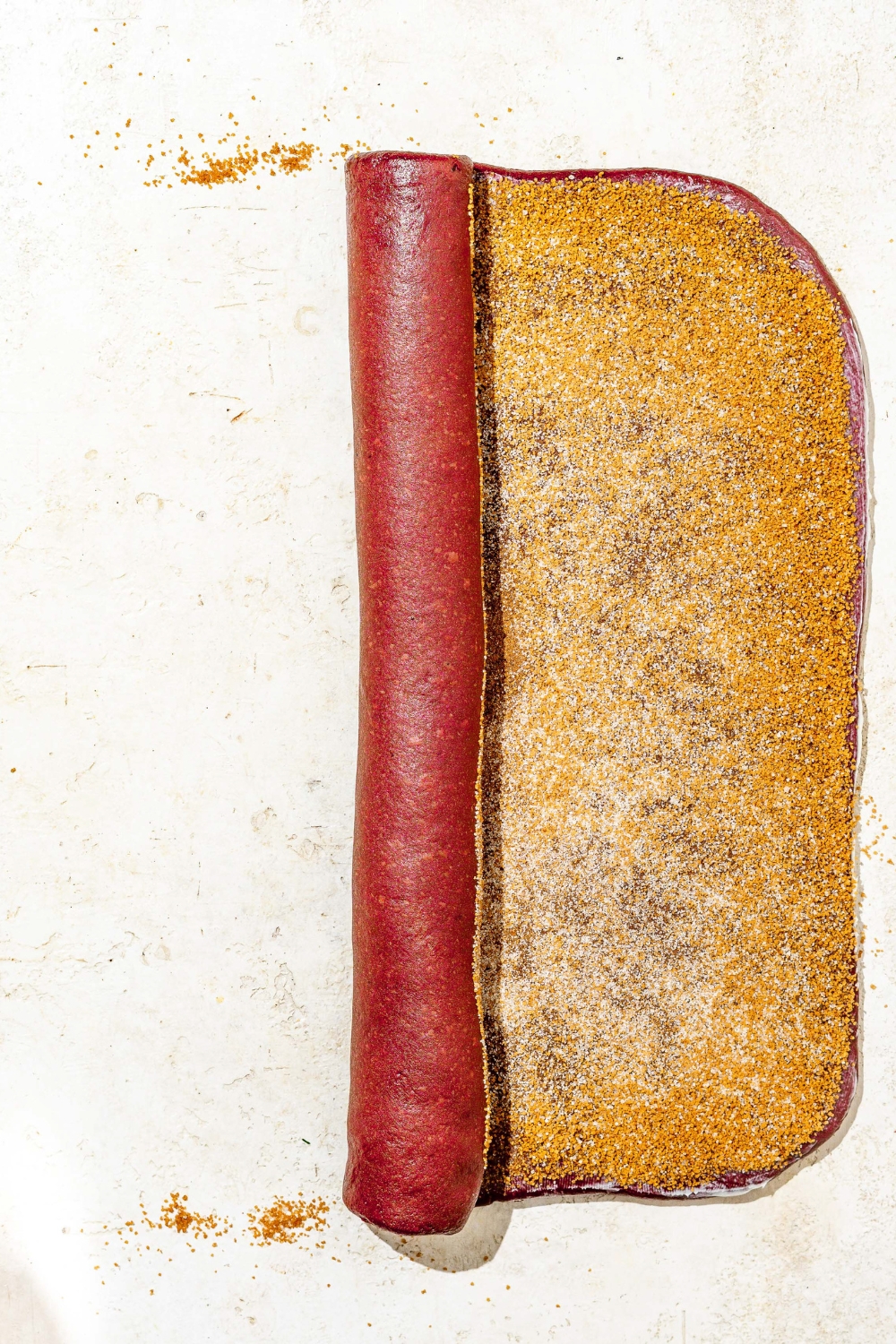 Red velvet cinnamon roll doll shaped into a rectangle topped with cinnamon sugar mixture. The dough is being rolled.