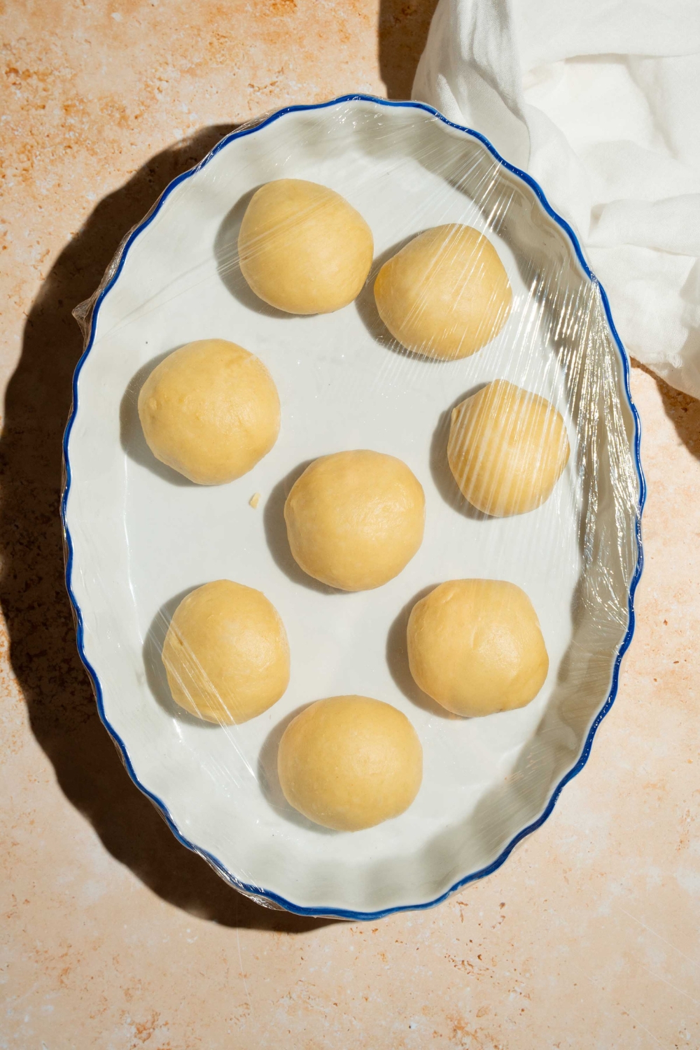 A baking dish with several balls of dinner roll dough. The dish is wrapped in saran wrap. The dish is on a tan counter with a white cloth napkin.