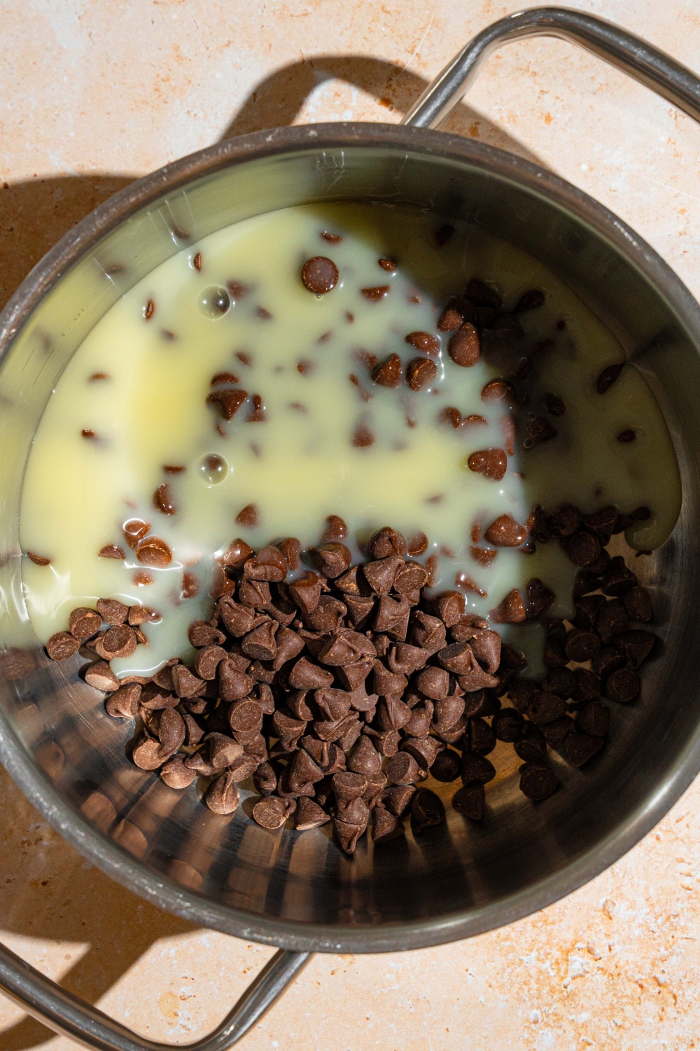 A stock pot with chocolate chips, condensed milk, and vanilla. The pot is on a tan counter.