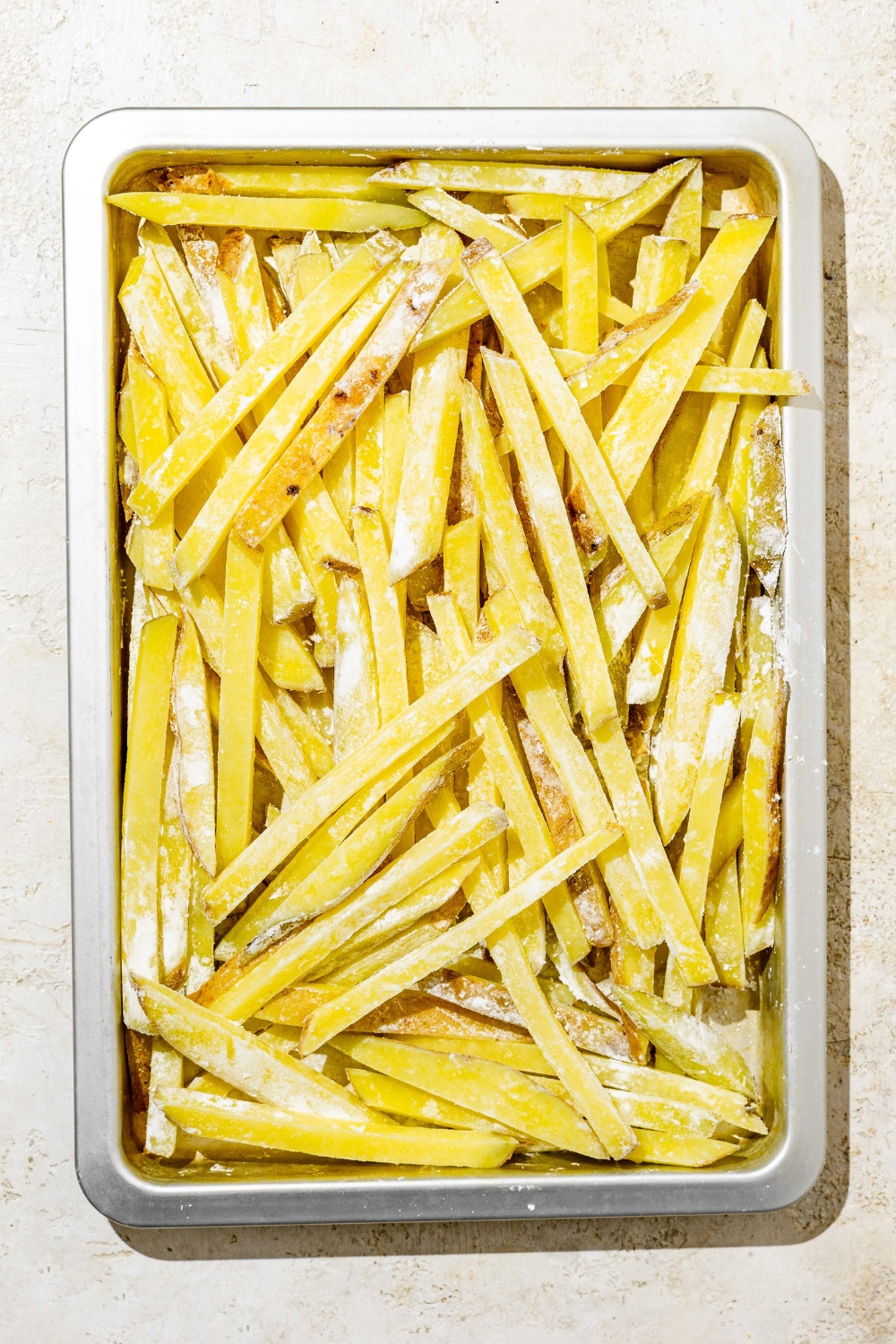 A baking sheet with boiled and drained french fries coated with corn starch. The sheet is on a white counter.