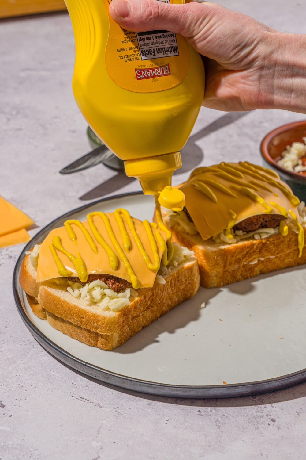 Two open grilled cheeseburgers on a white plate. The burgers are layered with shredded mozzarella, a beef patty, and a slice of American cheese with a hand squeezing mustard over the burgers.