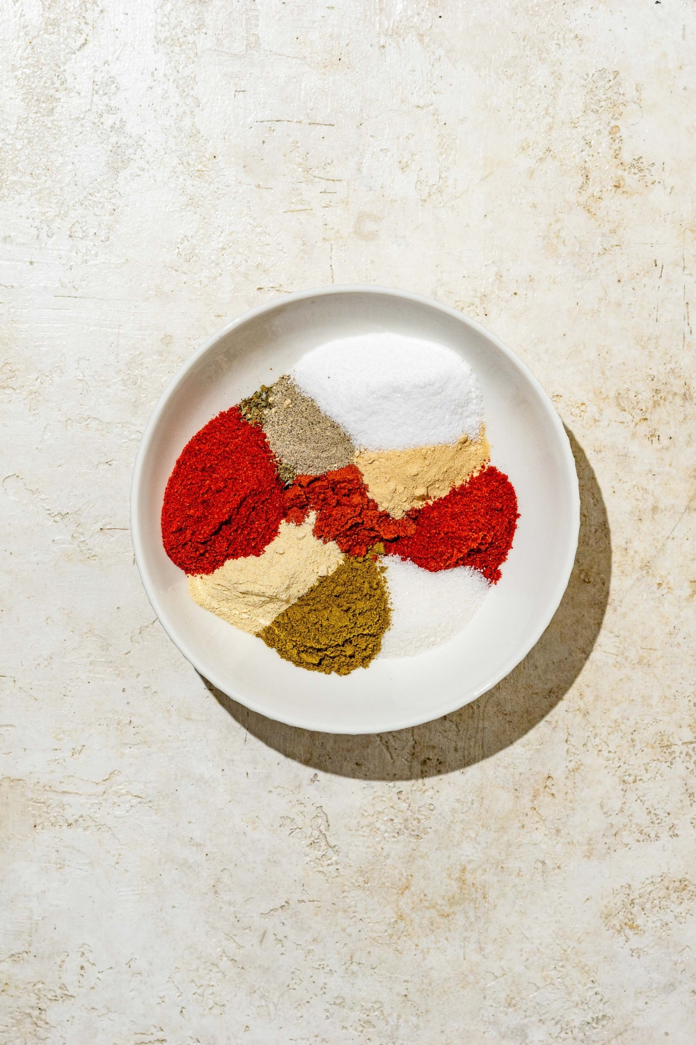 A white bowl with measured ingredients to make Red Robin seasoning including salt, paprika, onion powder, garlic powder, cumin, chili powder, and more. The bowl is on a tan counter.