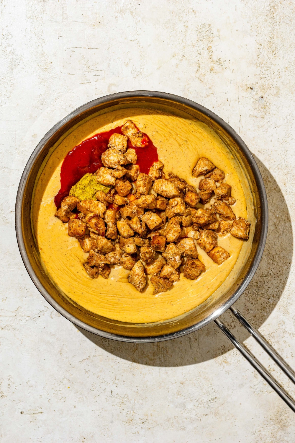 A skillet with Buffalo mac and cheese sauce topped with cooked seasoned chicken, hot sauce, and mustard. The skillet is on a tan counter.