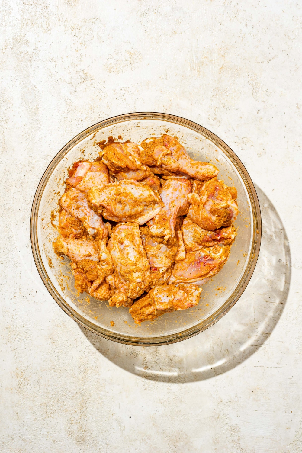 A glass bowl with uncooked chicken wings tossed in seasoning mixture. The bowl is on a tan counter.
