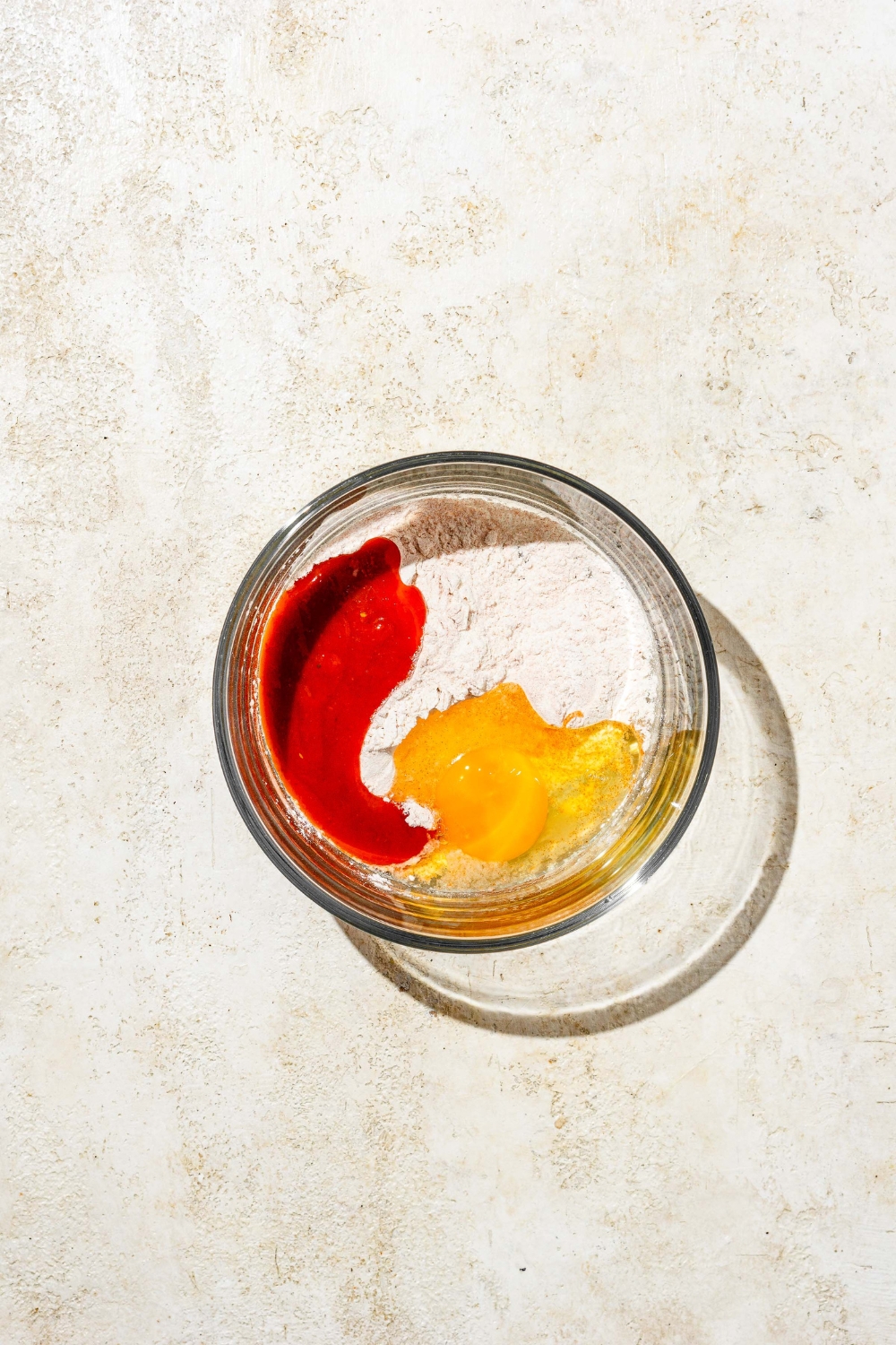 A glass bowl with ingredients to make a wet batter for hot honey chicken tenders including egg, hot sauce, and flour. The bowl is on a tan counter.