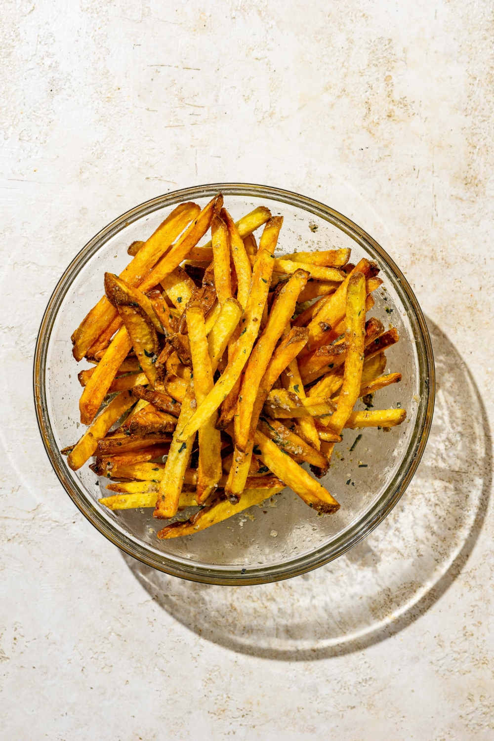 A glass bowl with cooked french fries tossed in garlic parmesan butter sauce. The bowl is on a white counter.