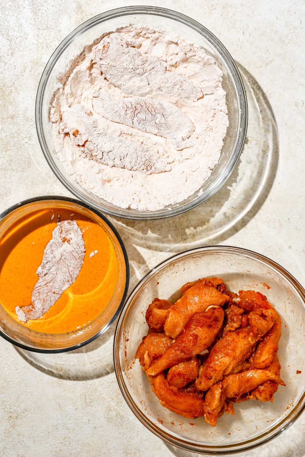 Three glass bowls with ingredients to bread chicken tenders including a bowl of seasoned chicken tenders, a bowl of flour mixture, and a bowl of wet batter.