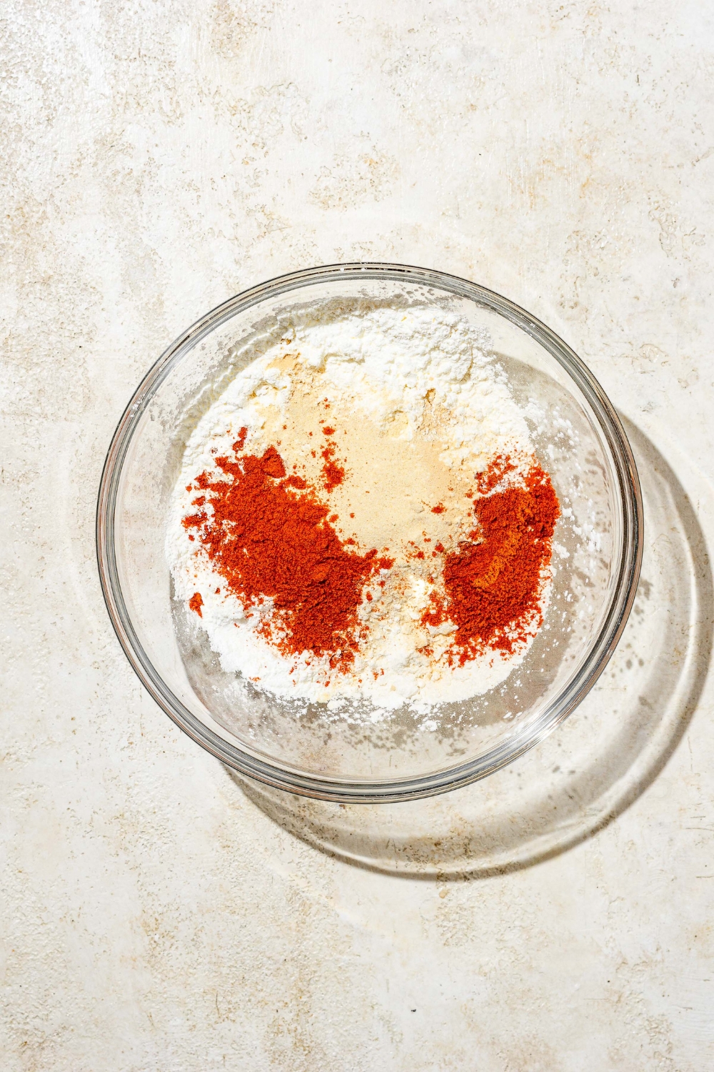 A glass bowl with ingredients to make hot honey flour mixture including flour, smoked paprika, onion powder, garlic powder, cornstarch, and more. The bowl is on a tan counter.