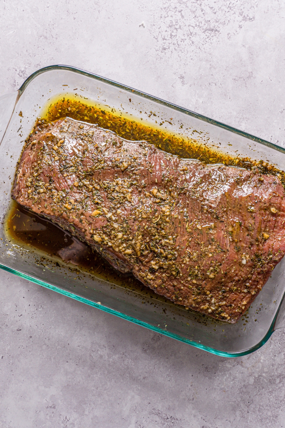 A whole top round steak sitting in a glass baking dish with covered with marinade. The dish is on a stone counter.