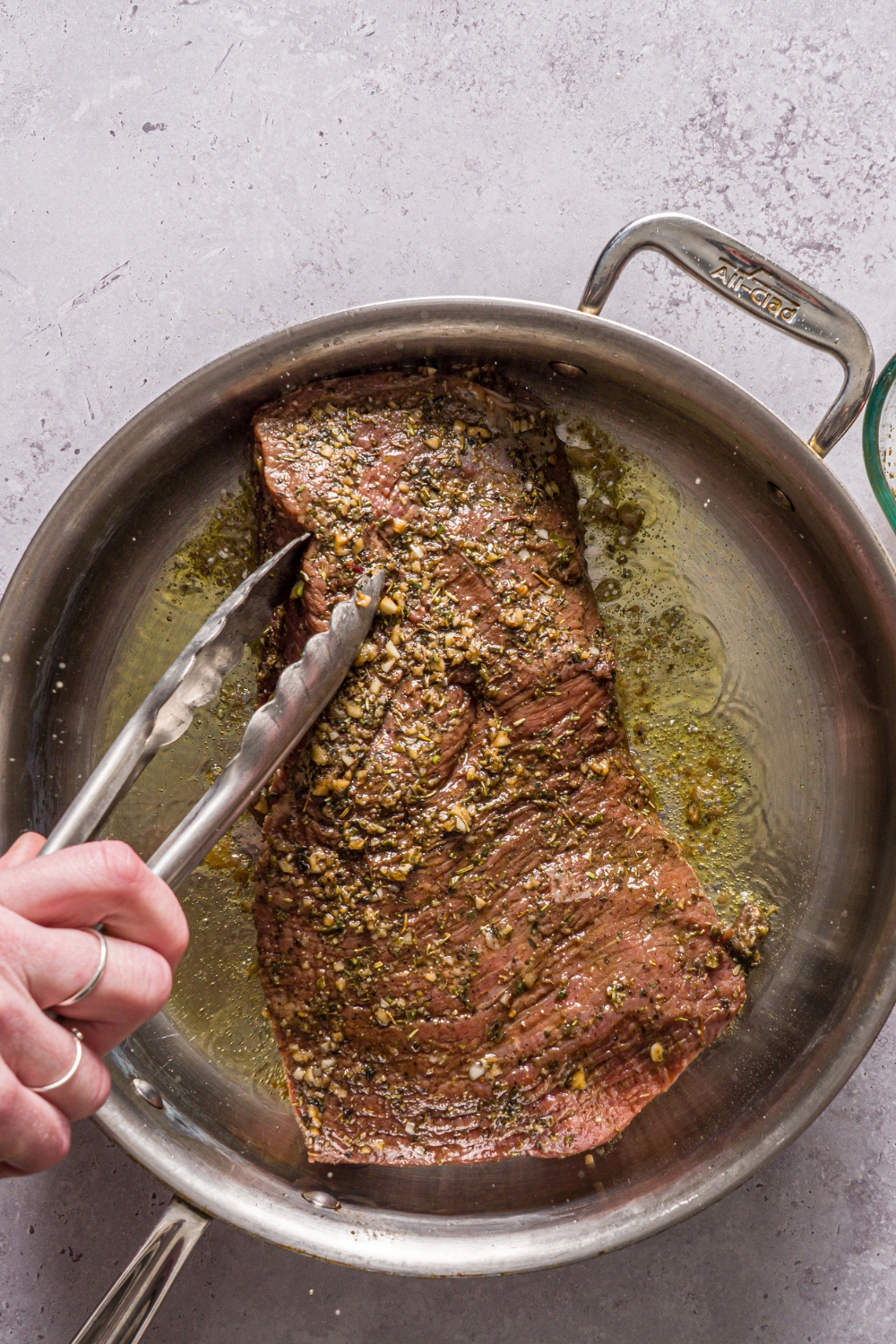A skillet with a marinated steak searing in oil. There is a pair of tongs flipping the steak. The skillet is on a stone counter.