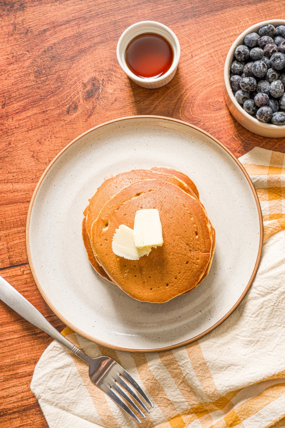 A white plate with a stack of muffin mix pancakes topped with butter. The plate is on a wooden counter with a small bowl of blueberries, cup of syrup, and yellow napkin.