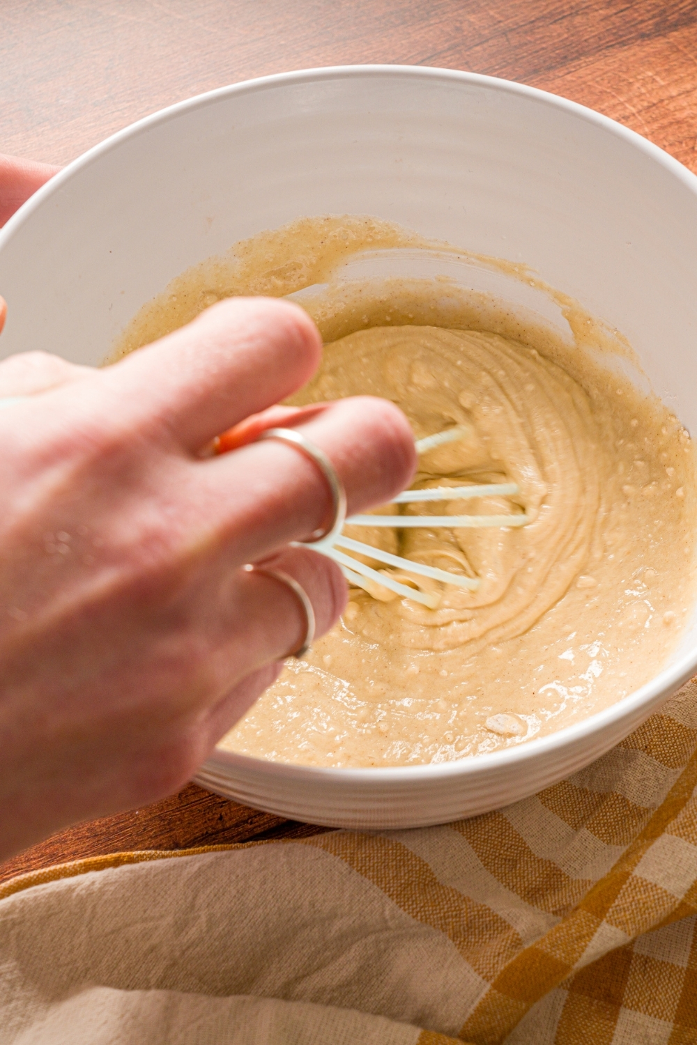 A hand whisking muffin mix pancake batter in a white bowl. The bowl is on a wooden counter with a yellow striped napkin.
