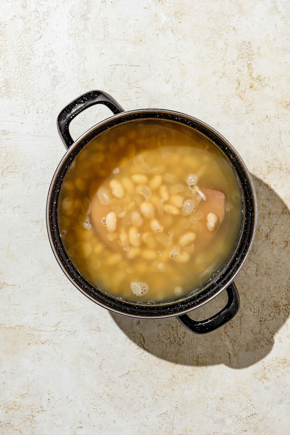 A stock pot with beans and ham cooking in water. The pot is on a tan counter.