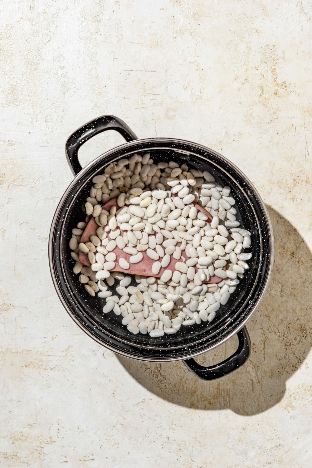 A stock pot with navy beans and ham. The pot is on a tan counter.