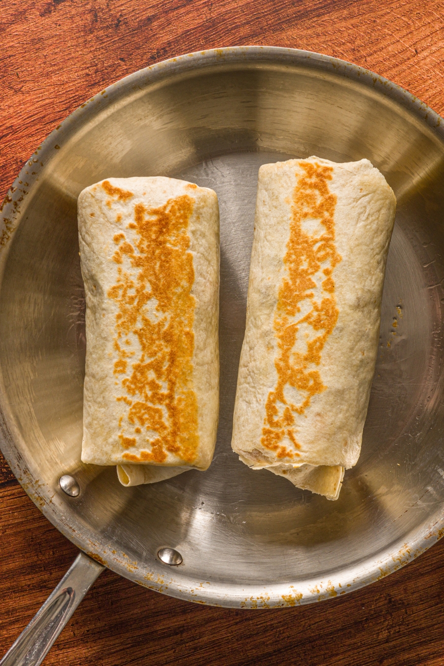 A skillet with two BBQ chicken wraps searing in oil. The skillet is on a wooden counter.