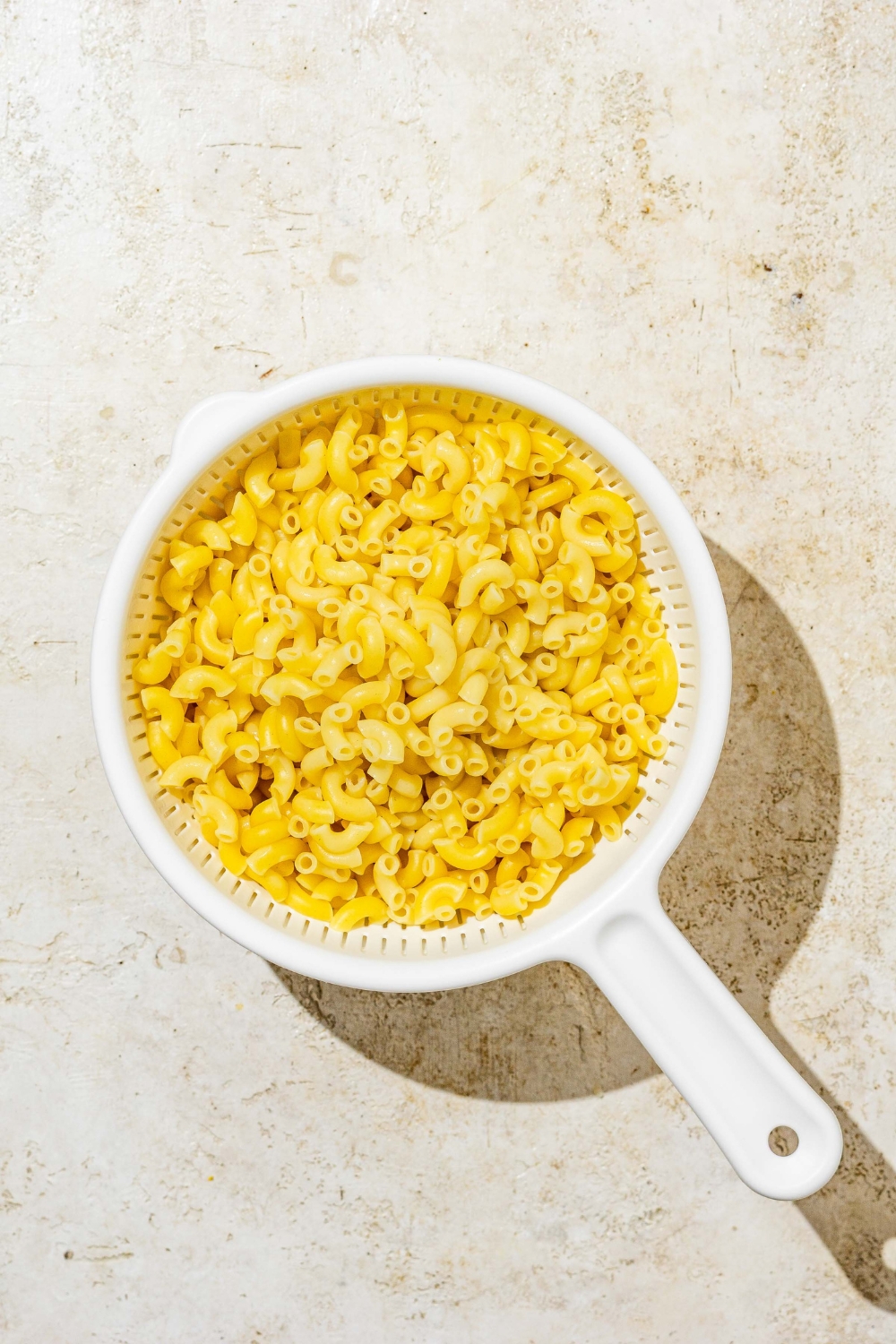 A white colander with cooked elbow pasta. The colander is on a tan counter.
