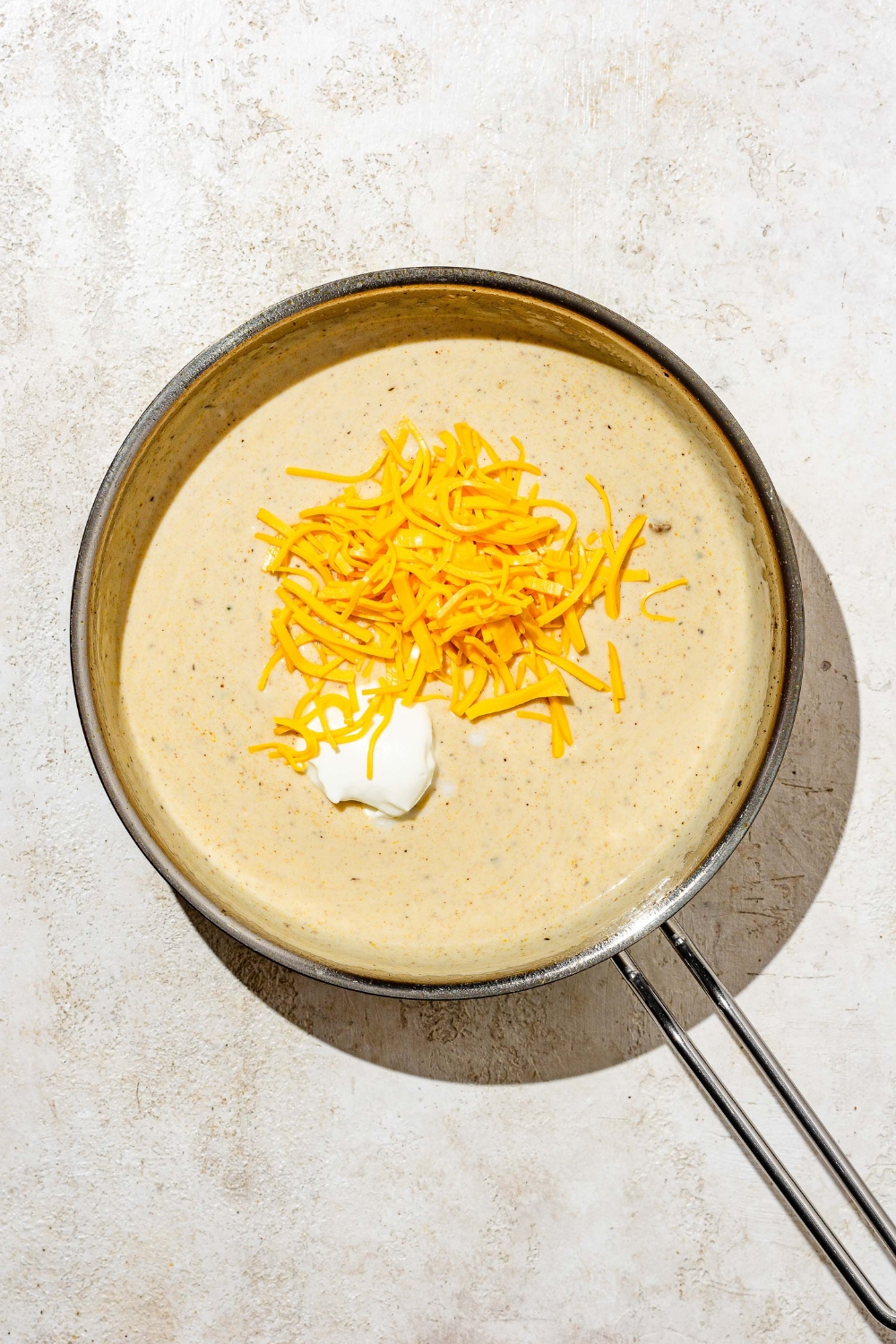 A skillet with a creamy cheese sauce topped with shredded cheddar cheese and boursin cheese. The skillet is on a tan counter.