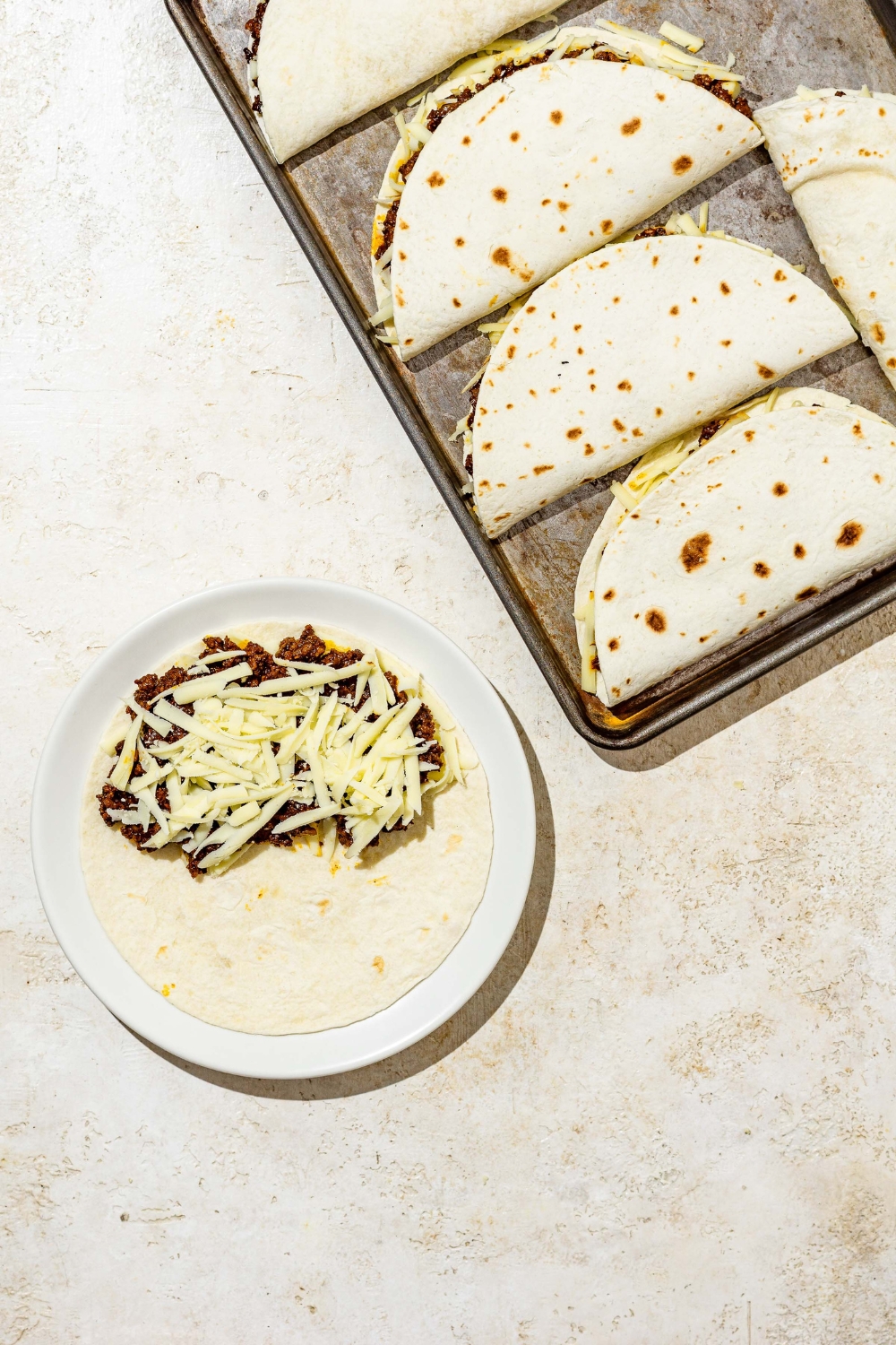 A white plate with an open taco layered with beef and shredded mozzarella cheese on a tan counter with a baking sheet of tacos.