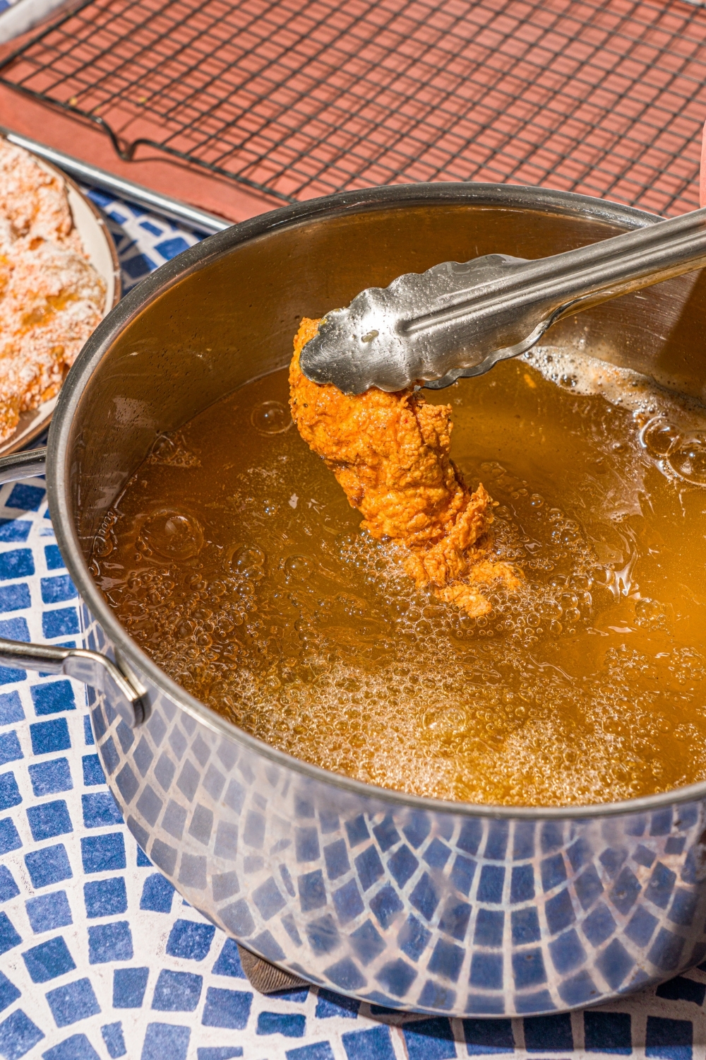 A pot of frying oil with tongs holding a breaded fried chicken tender. The pot is on a blue tiled counter.