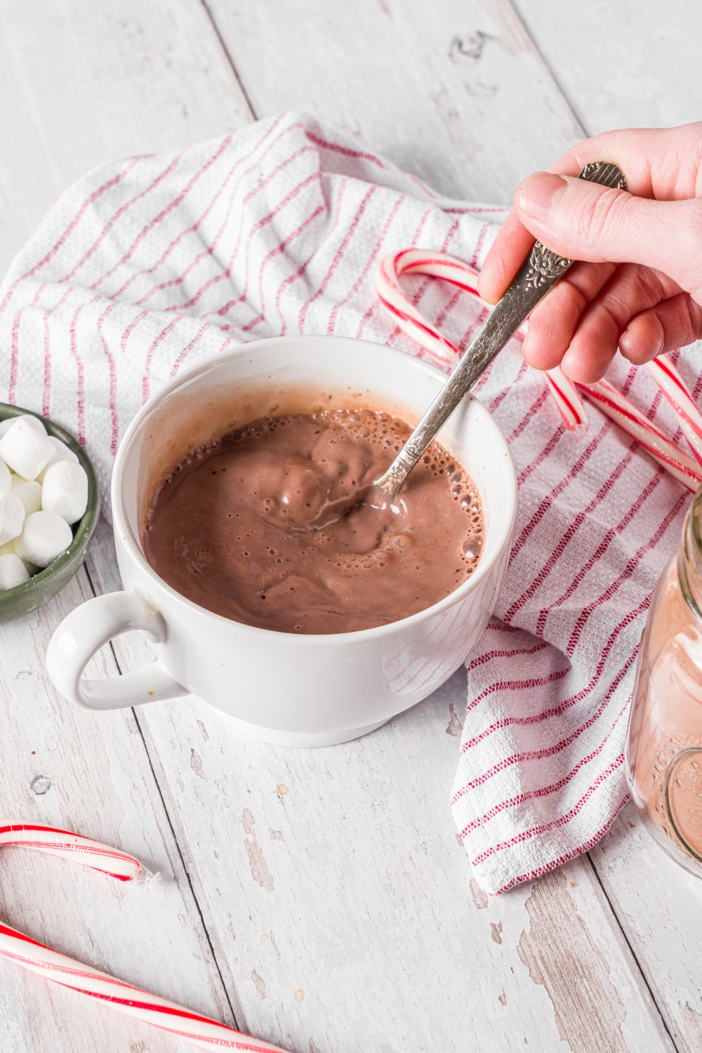 A white mug with homemade hot cocoa with a hand stirring the cocoa with a spoon. The mug is on a wooden counter with a red striped napkin and candy canes.