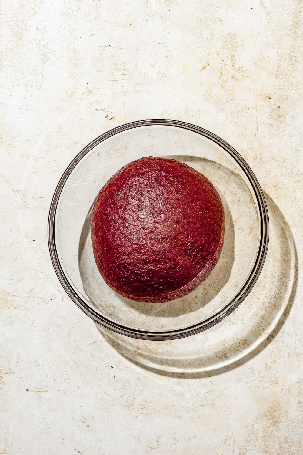 A glass bowl with a ball of red velvet cinnamon roll dough. The bowl is on a tan counter.