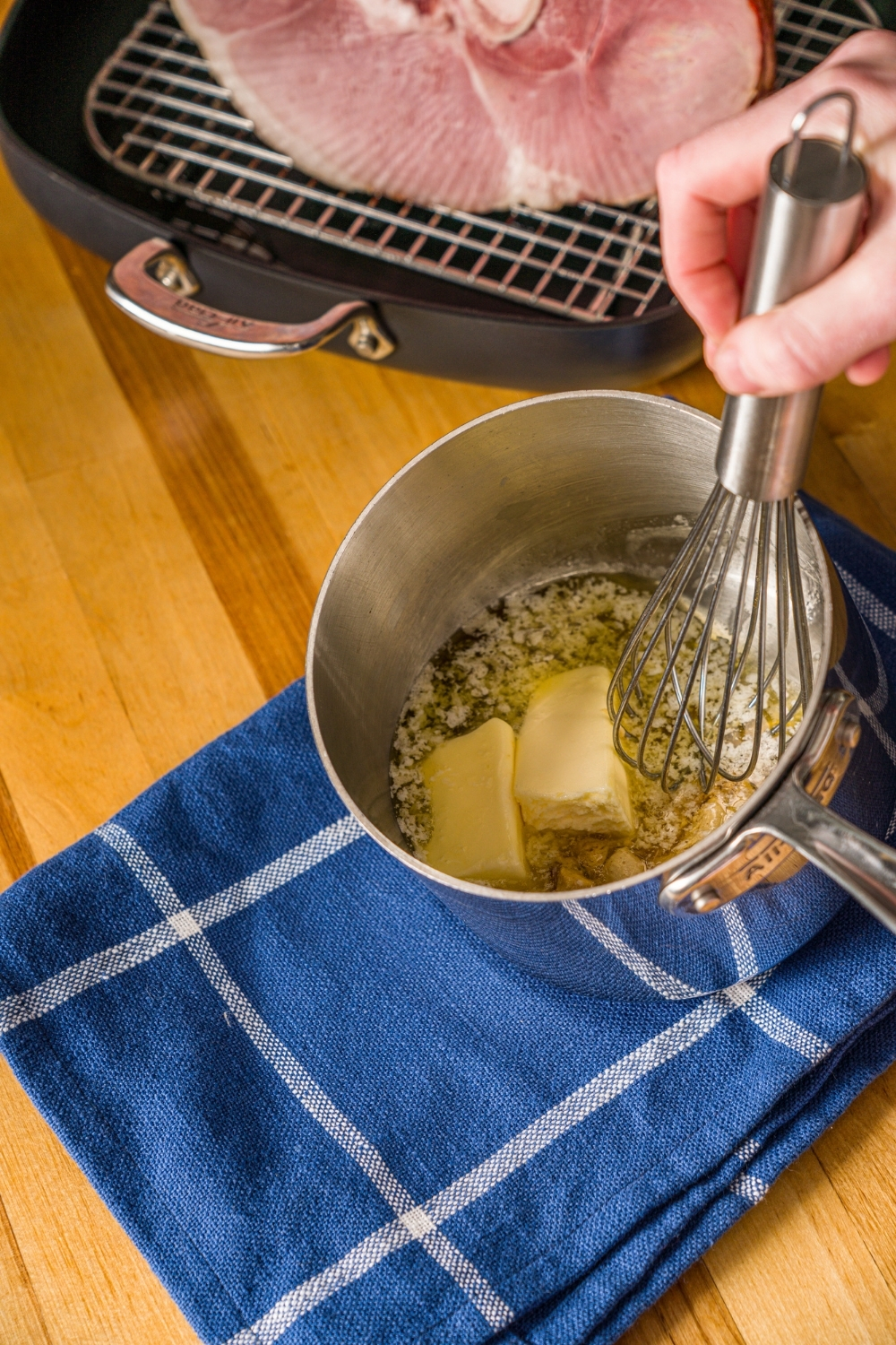 A sauce pan with a whisk mixing ingredients for spiral ham including melted butter, honey, and mustard. The pan is on a wooden counter with a blue cloth napkin and whole spiral ham in a roasting pan.