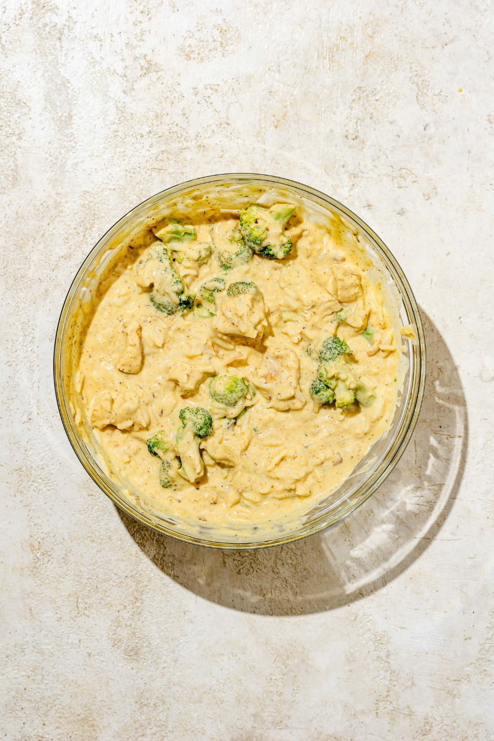 A glass bowl with broccoli florets, chicken, and rice mixed with a creamy cheese sauce. The bowl is on a tan counter.