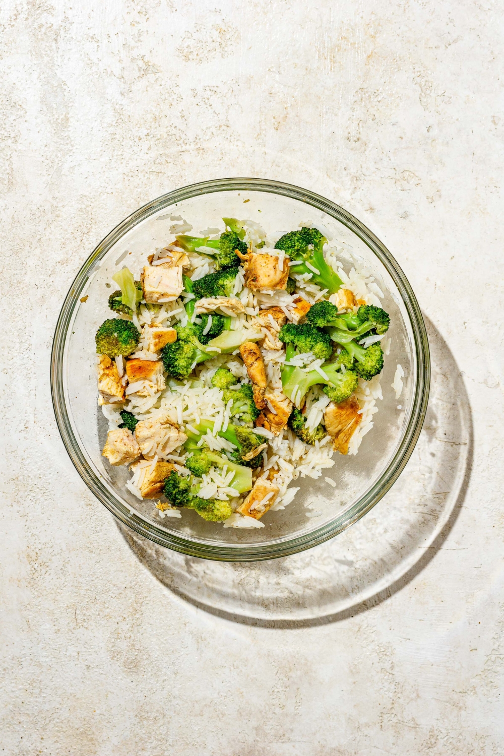A glass bowl with cubed chicken, broccoli florets, and rice. The bowl is on a tan counter.