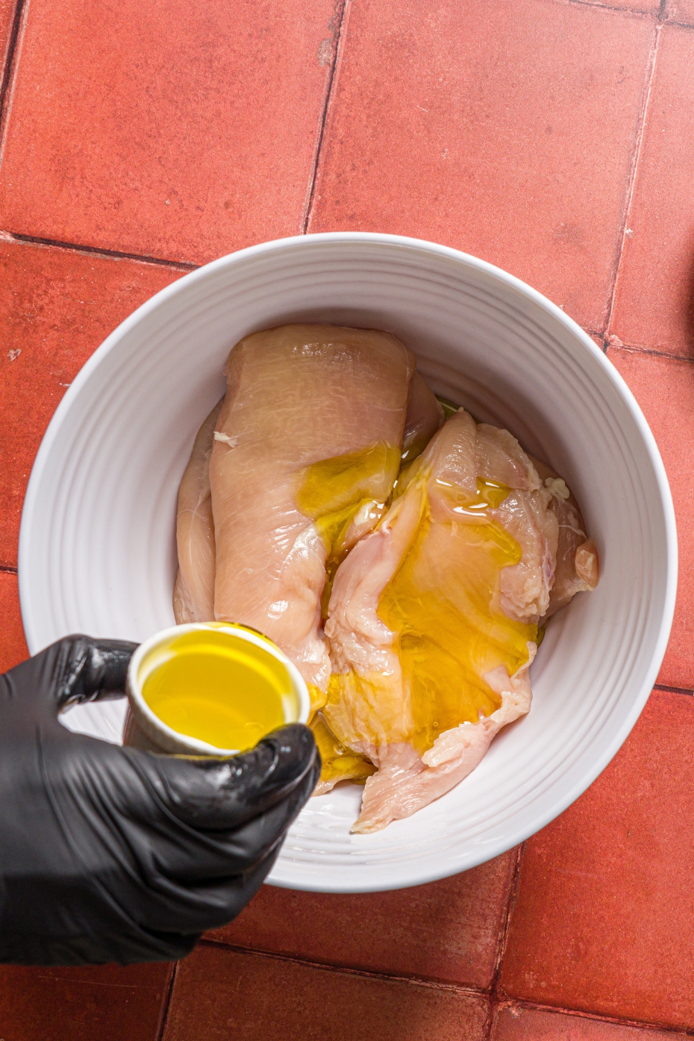 A white bowl with uncooked thin sliced chicken breasts with a hand pouring olive oil from a small bowl over the chicken.