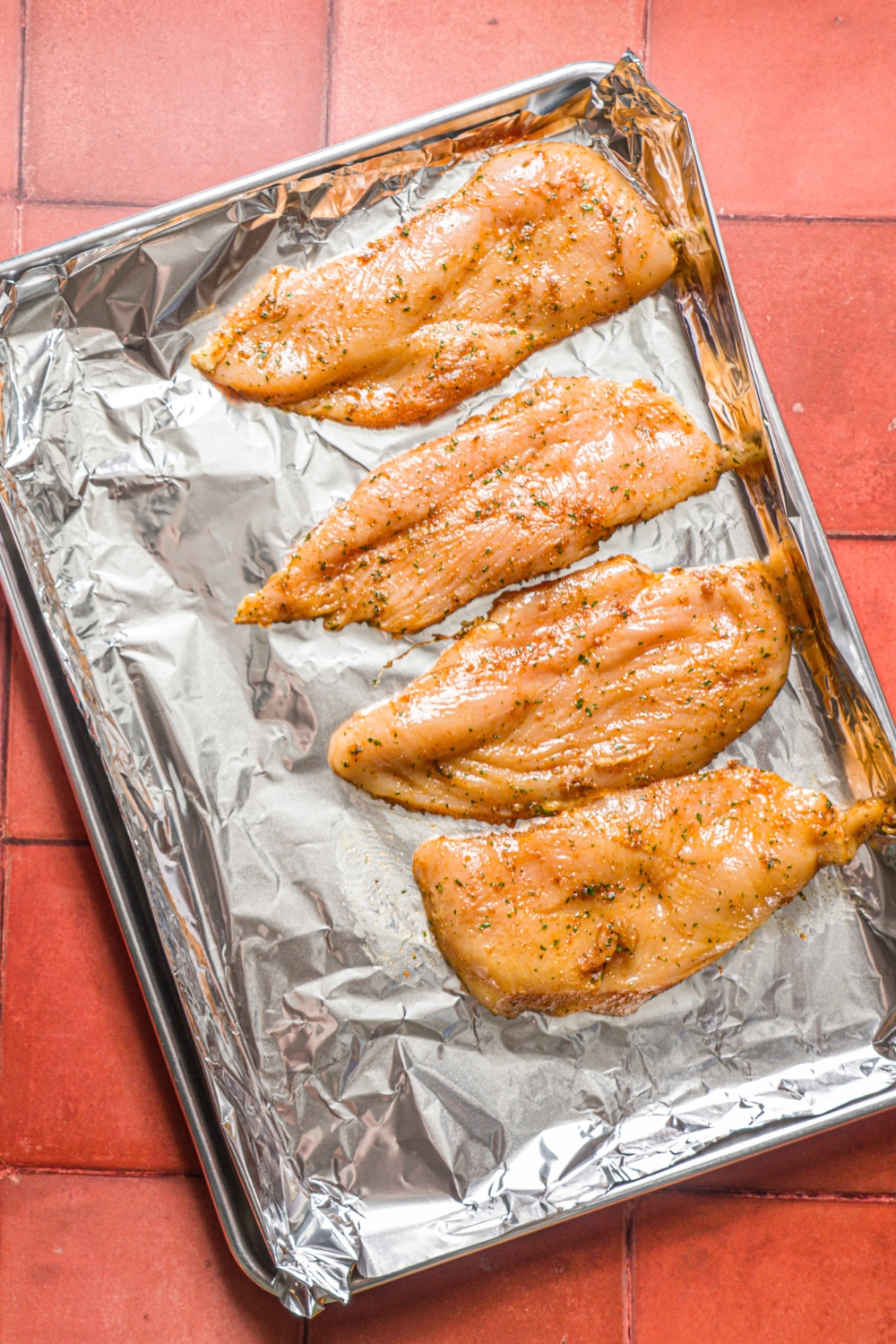A baking sheet lined with aluminum foil with four pieces of thin sliced uncooked chicken breasts coated in oil and seasonings. The sheet is on a red tiled counter.