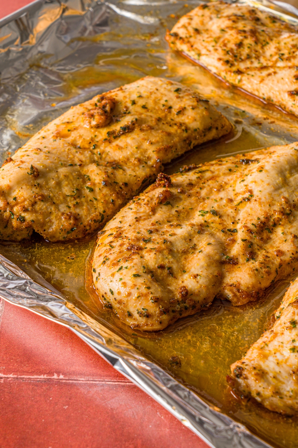 A baking sheet lined with aluminum foil with several pieces of baked thin sliced seasoned chicken breasts. The baking sheet is on a red tiled counter.