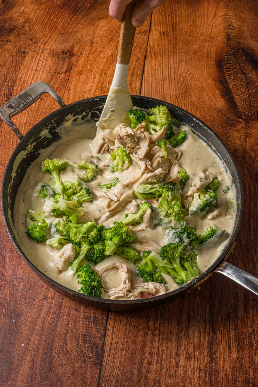 A skillet with steamed broccoli and shredded turkey tossed in turkey divan sauce. A spatula is stirring the mixture. The skillet is on a wooden counter.