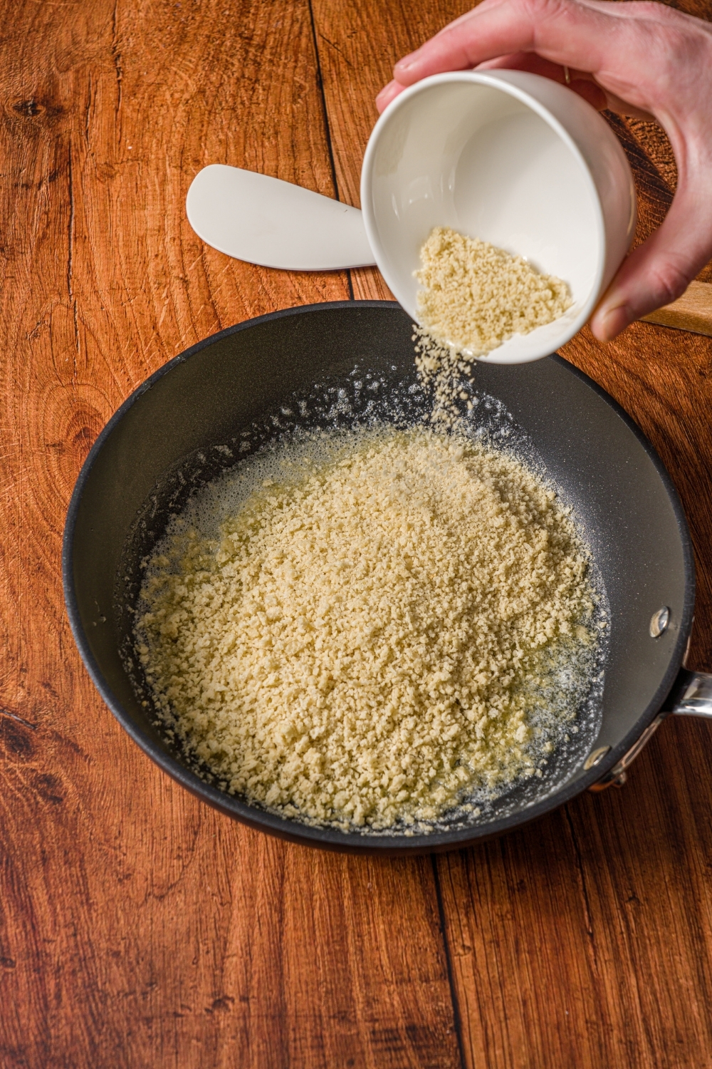 A small skillet with panko breadcrumbs cooking in butter. A hand is dumping a small bowl of panko into the skillet.