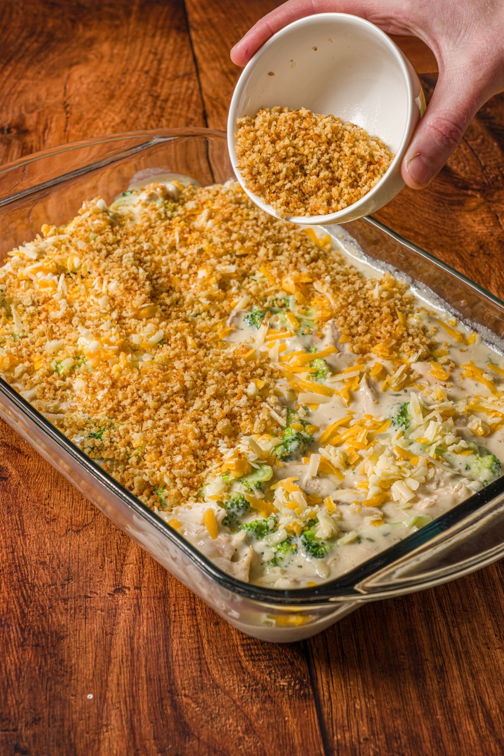 A baking dish with uncooked turkey divan with a bowl topping the mixture with toasted panko breadcrumbs. The dish is on a wooden counter.