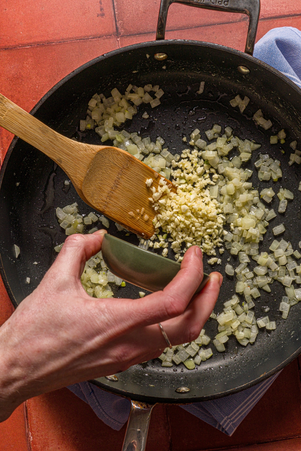 A skillet with diced onion cooking in oil. There is a hand pouring garlic into the skillet with a wooden spoon stirring the mixture.