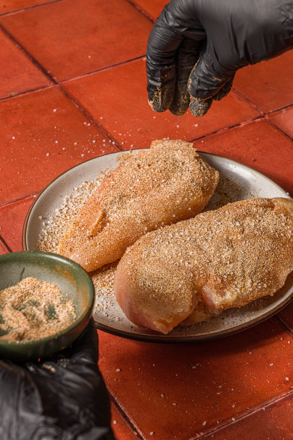 Two uncooked chicken breasts on a white plate on a tiled counter. There is a hand sprinkling a seasoning mixture over the chicken.