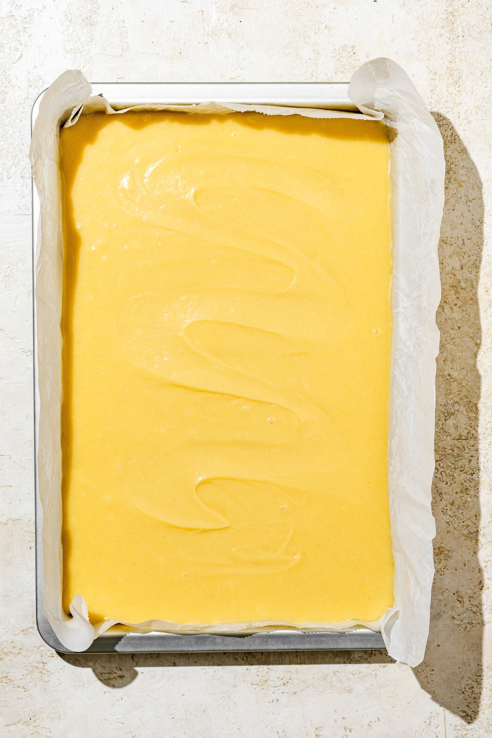 A baking pan lined with parchment paper with uncooked white Texas sheet cake patter in the pan. The sheet is on a white counter.