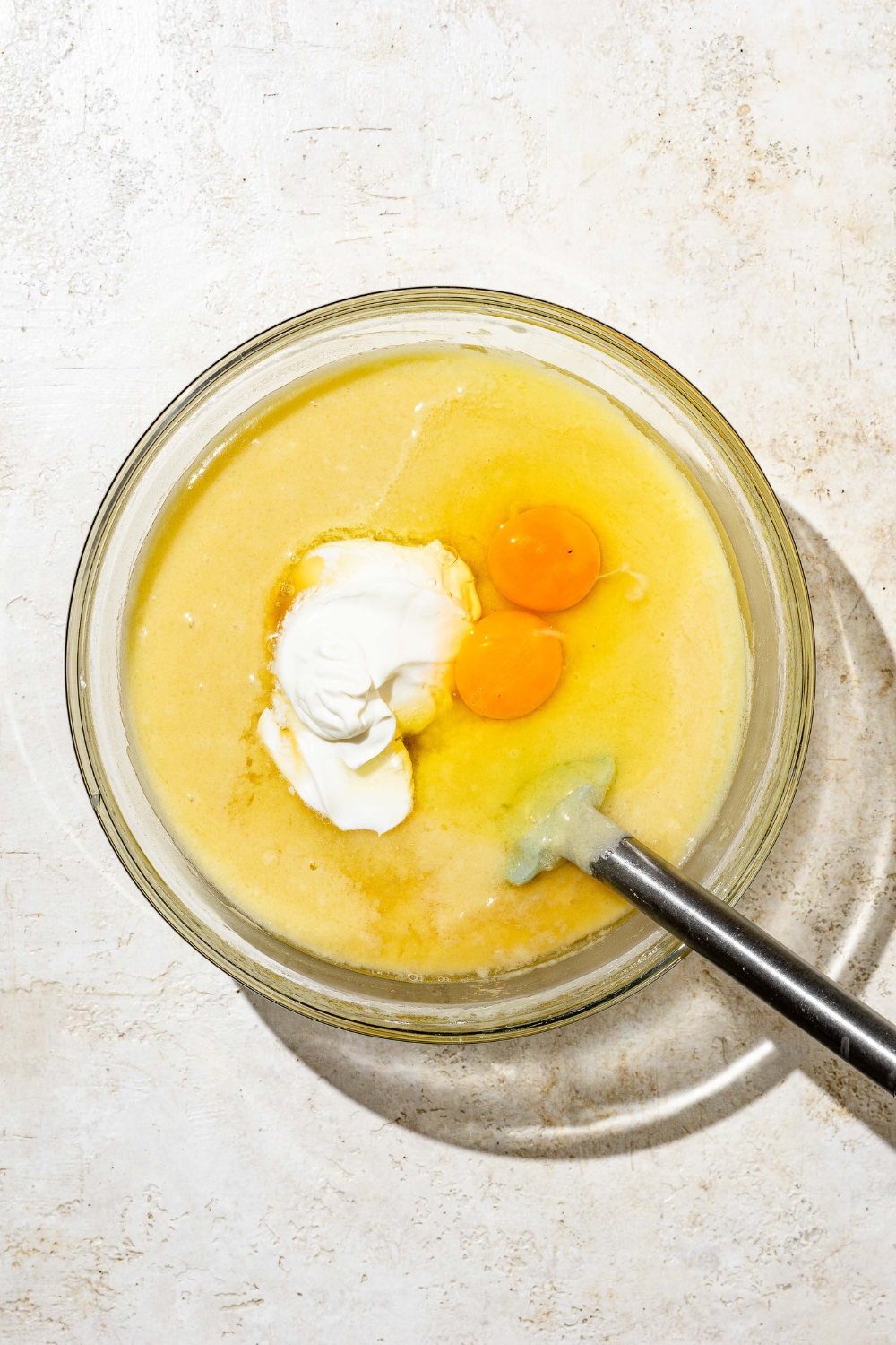 A glass bowl with wet ingredients combining with dry ingredients to make white Texas sheet cake batter. The batter is topped with eggs and sour cream. There is a spatula in the bowl. The bowl is on a white counter.