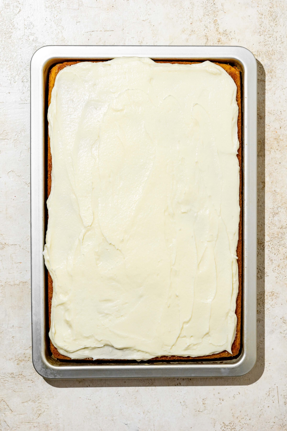A baking pan with baked white Texas sheet cake topped with a layer of icing. The pan is on a white counter.