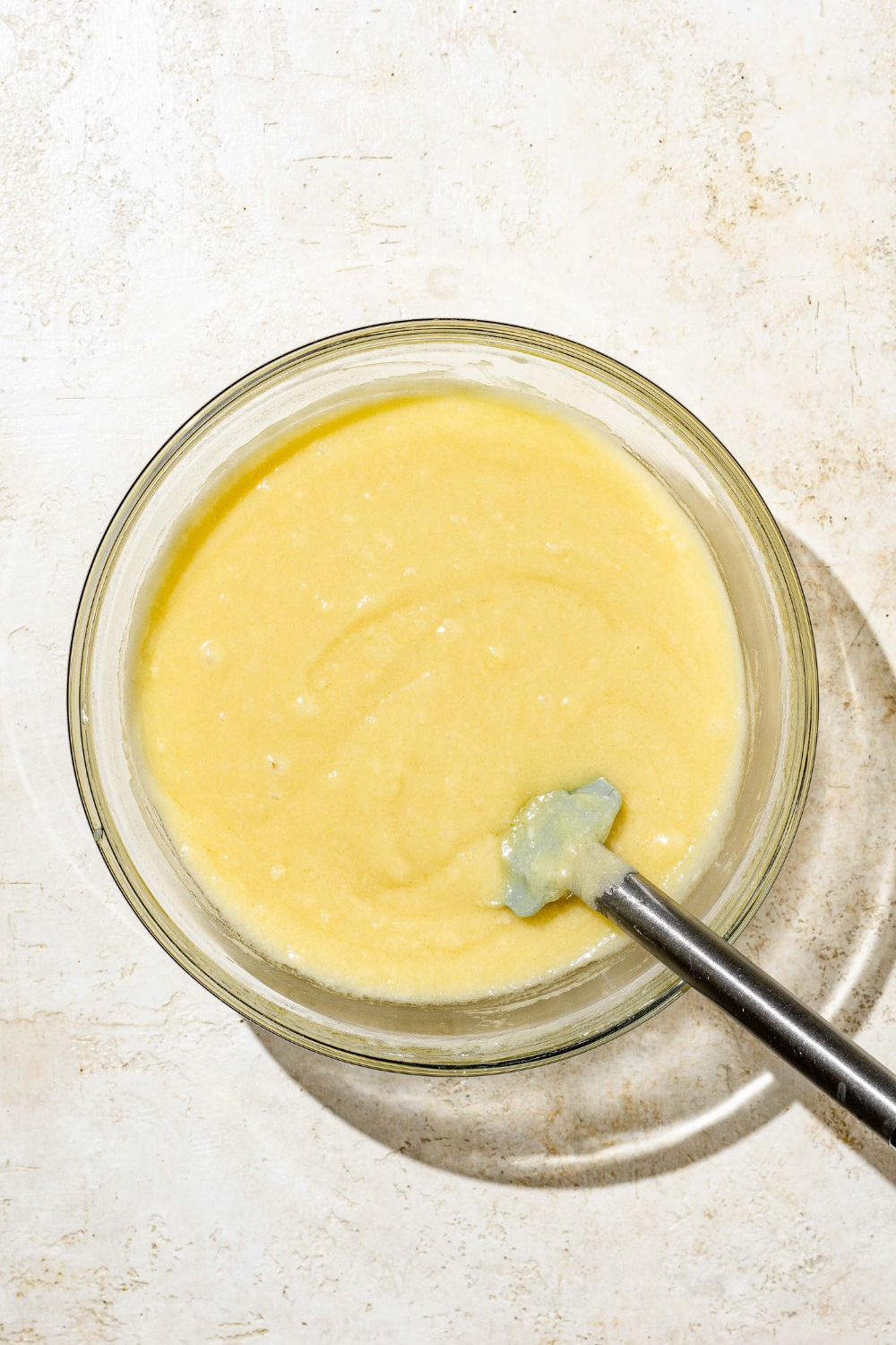 A glass bowl with a spatula combining wet and dry ingredients for white Texas sheet cake batter. There is a spatula in the bowl. The bowl is on a white counter.