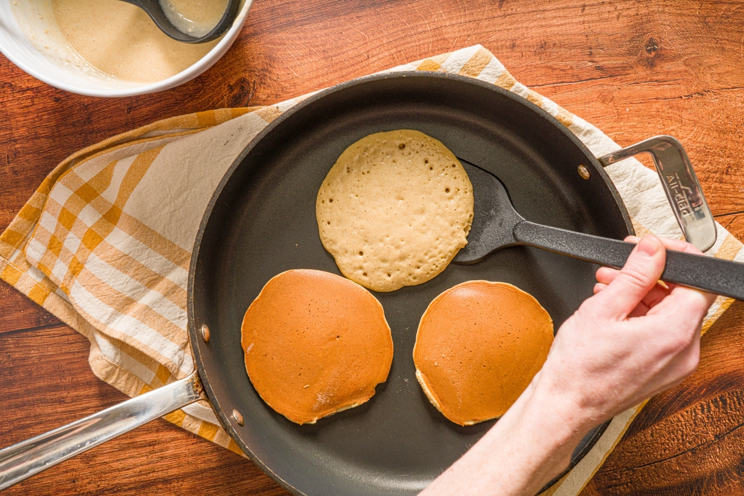 Three muffin mix pancakes cooking on a skillet with a spatula flipping one of the pancakes. The skillet is on a wooden counter with a yellow striped napkin.