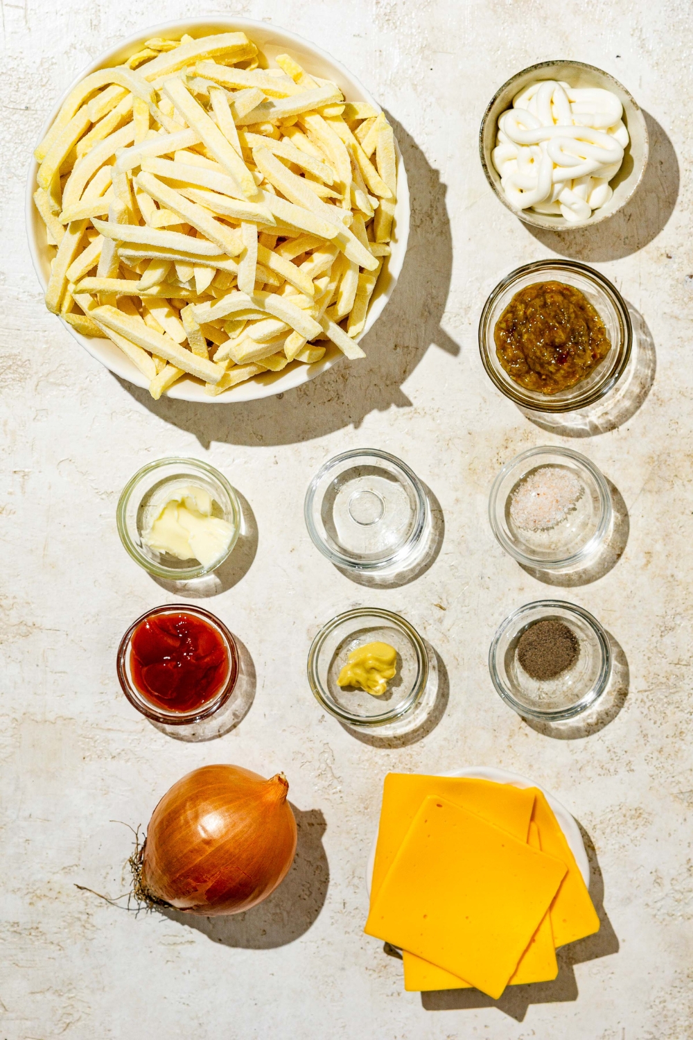 An overhead shot of several bowls in various sizes containing ingredients to make In n Out animal style fries including frozen fries, slices of American cheese, onion, ketchup, mayo, mustard, oil, and seasonings.